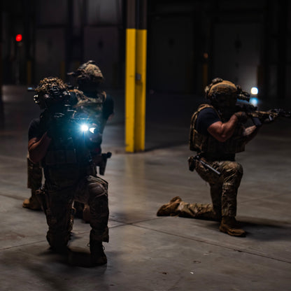 Two soldiers in tactical gear in a dark indoor setting with one holding a flashlight.